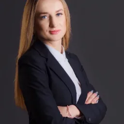 Portret kobiety w czarnym garniturze i białej koszuli, z blond włosami i niebieskimi oczami, pozującej na ciemnoszarym tle z założonymi rękami, na których widać delikatną bransoletkę.