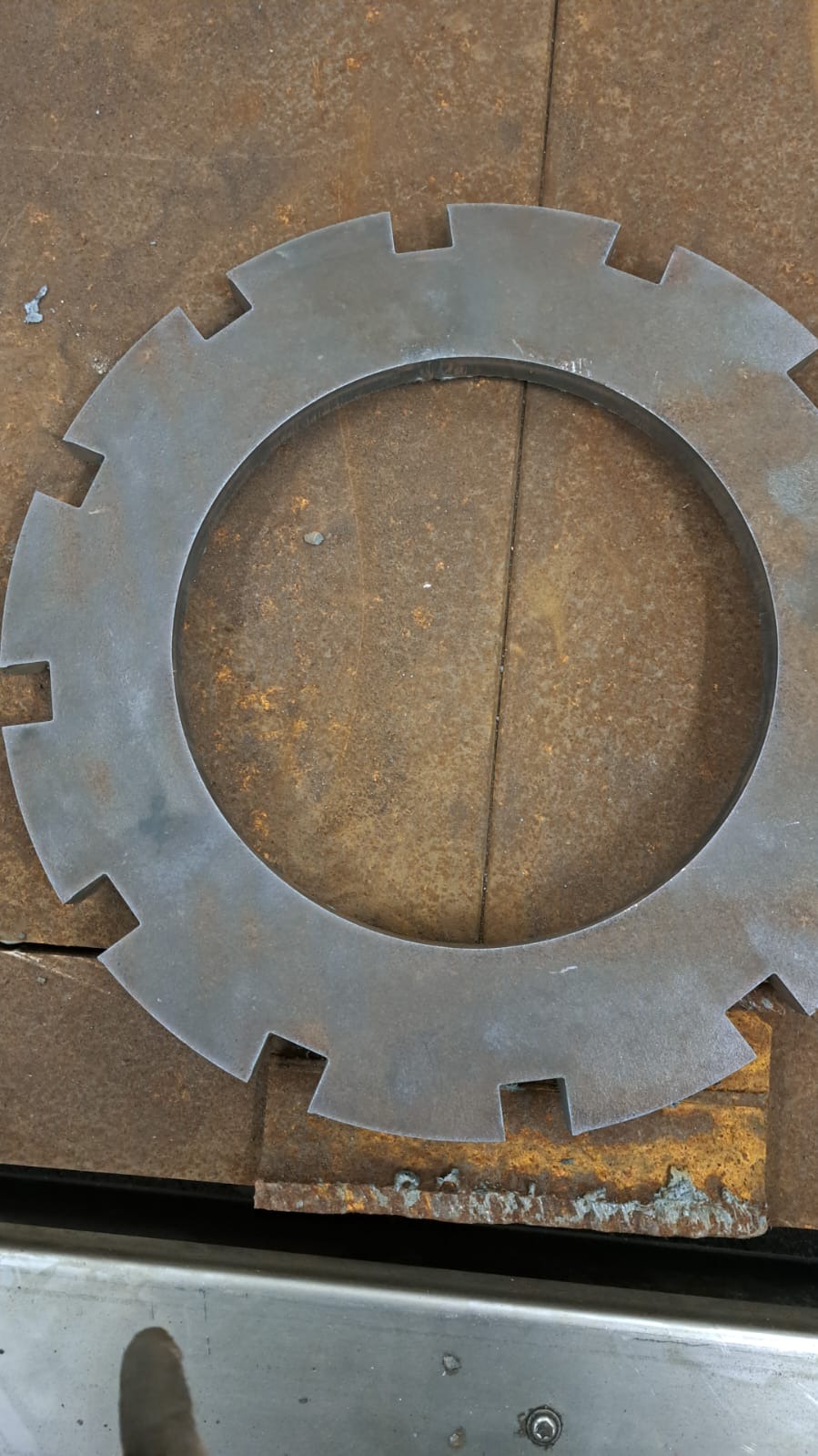 Wycięty metalowy element w kształcie koła z zębami na zardzewiałym stole warsztatowym. Widoczne ślady obróbki i niedoskonałości powierzchni.