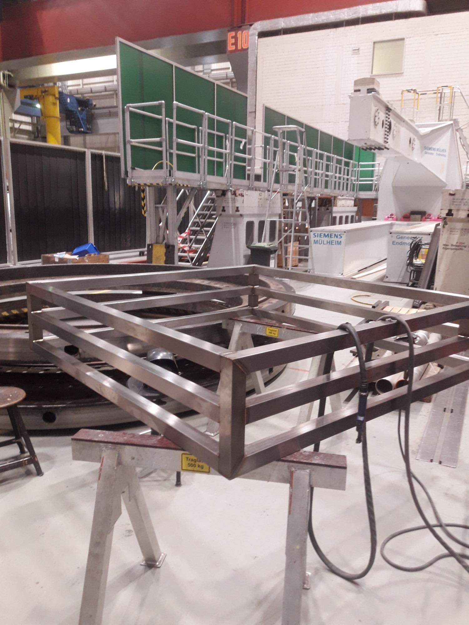 Metalowa rama konstrukcyjna na kozłach w hali przemysłowej, widoczne spawane łączenia i przewody spawalnicze, w tle elementy infrastruktury przemysłowej.