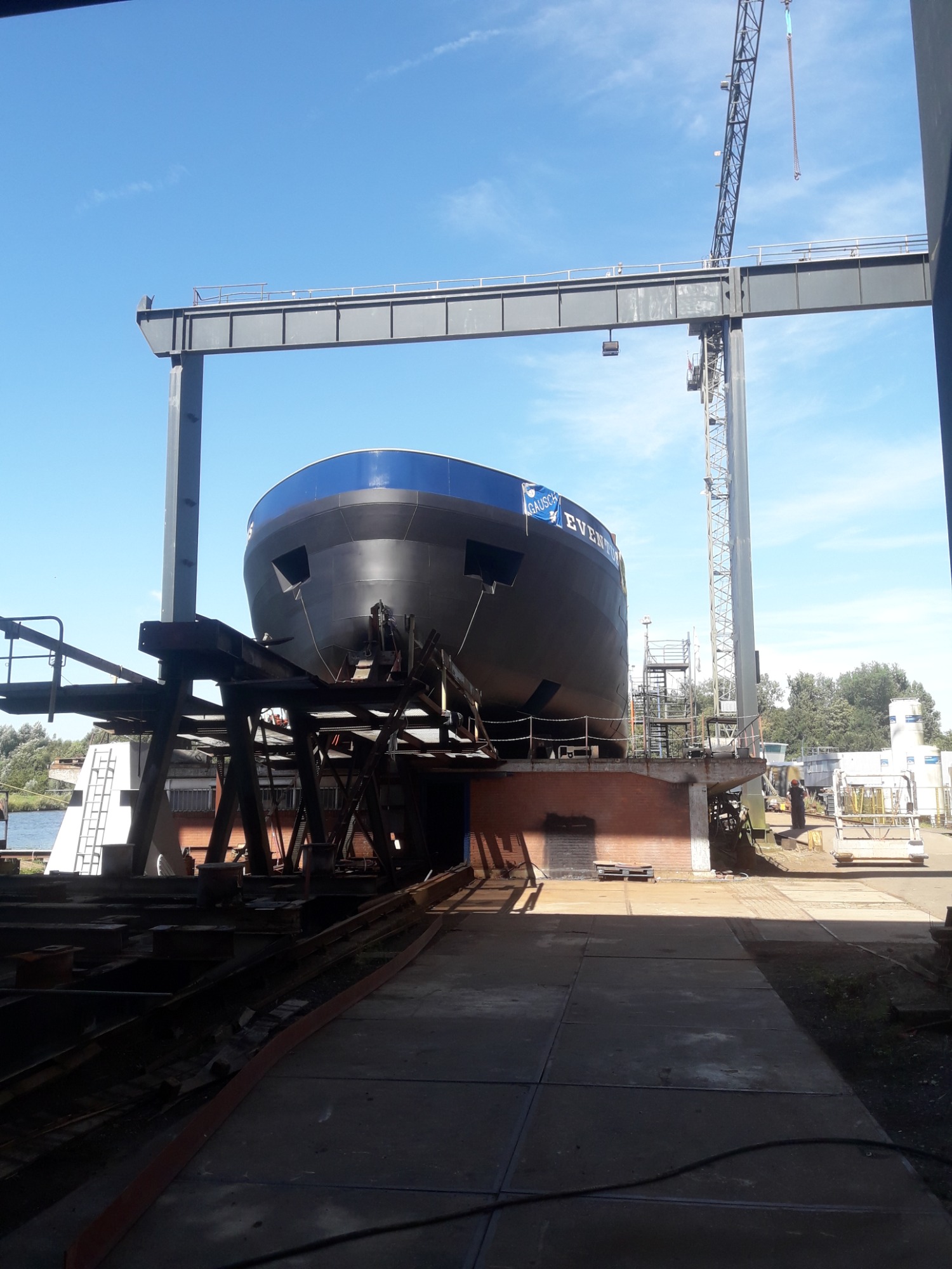 Kadłub statku w doku, pod suwnicą bramową, widoczne elementy konstrukcyjne i rusztowania, etap budowy.