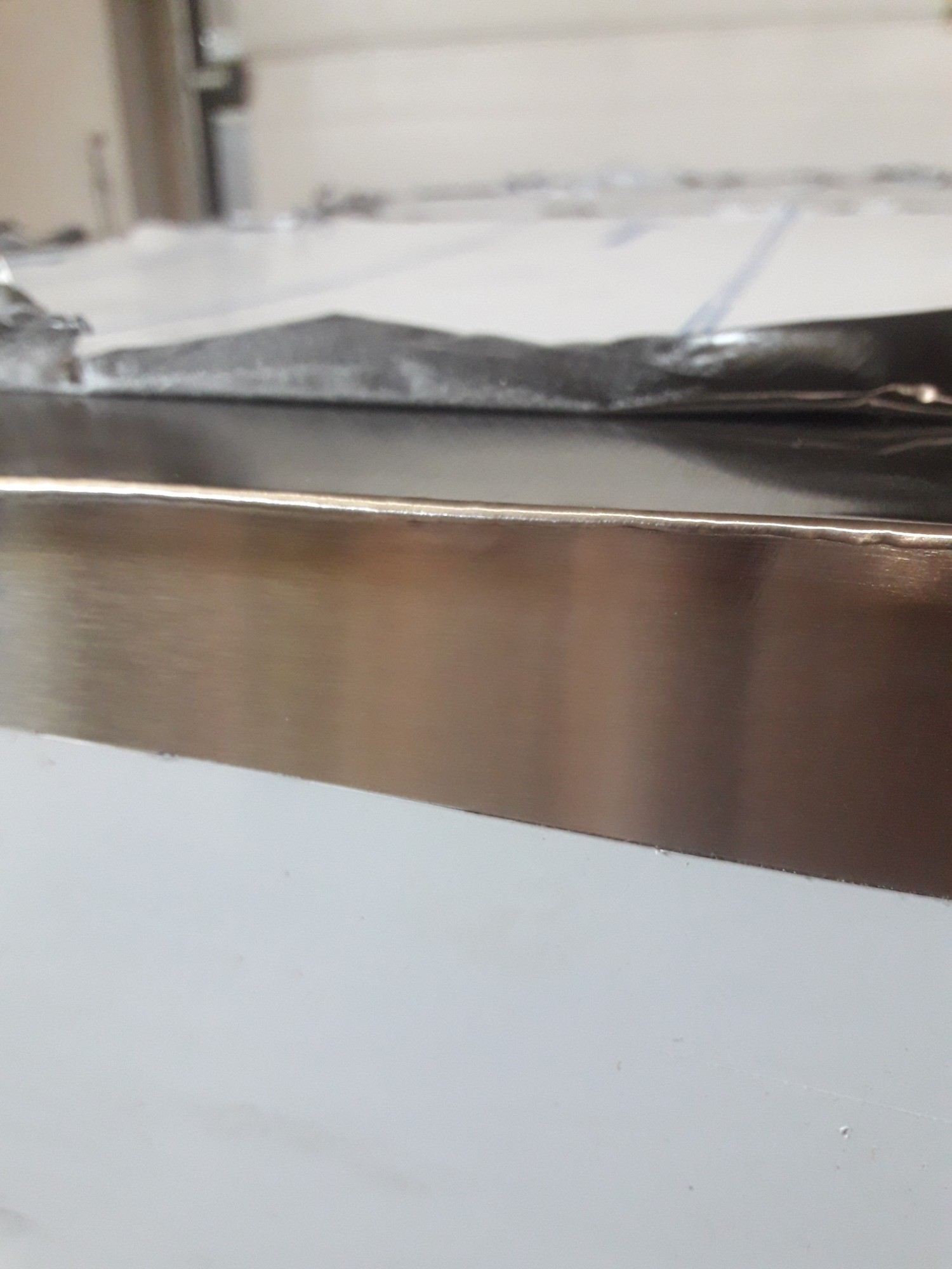 Metalowa listwa ochronna, prawdopodobnie ze stali nierdzewnej, zamontowana na białej powierzchni, widoczne ślady folii ochronnej.