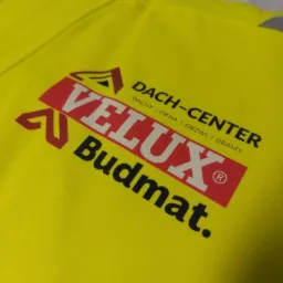 Żółta odzież robocza z nadrukowanym logo firmy Velux Budmat Dach-Center specjalizującej się w dachach, oknach, drzwiach i bramach.