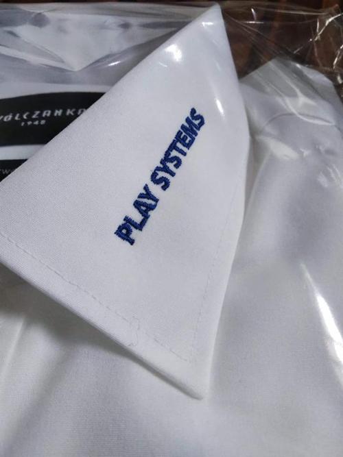Biała koszula z wyhaftowanym niebieskim napisem 'PLAY SYSTEMS' na kołnierzyku, widoczna część metki marki 'Wólczanka 1948', zapakowana w folię.