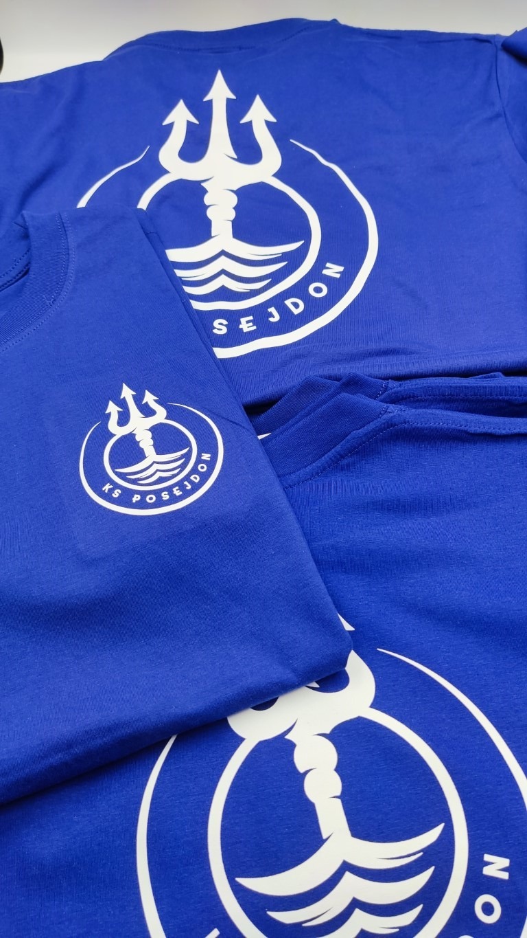 Stos niebieskich koszulek z białym nadrukiem logo KS Posejdon: trójząb z falami w okręgu z napisem.