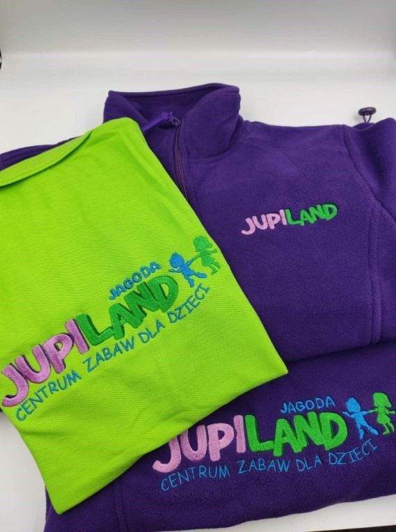 Zielona koszulka polo i fioletowy polar z wyhaftowanym kolorowym logo 'Jupiland Jagoda Centrum Zabaw dla Dzieci'.