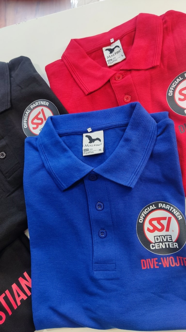 Trzy złożone koszulki polo w kolorach czarnym, niebieskim i czerwonym, z nadrukowanym logo SSI Dive Center i nazwą Dive-Wojtek, widoczne metki marki Malfini.