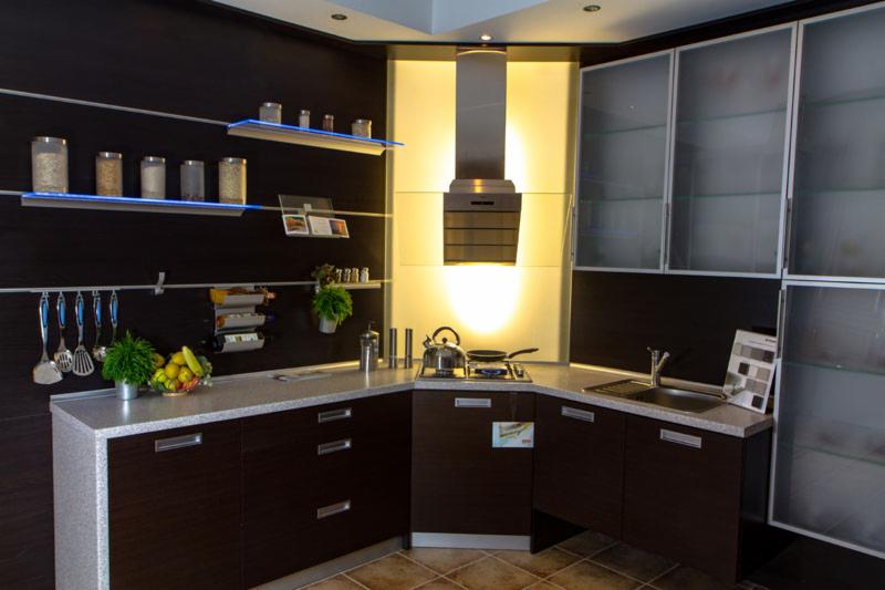 Nowoczesna kuchnia z ciemnymi frontami, jasnym blatem i oświetleniem LED pod półkami, wyposażona w srebrny okap i przeszklone szafki górne.