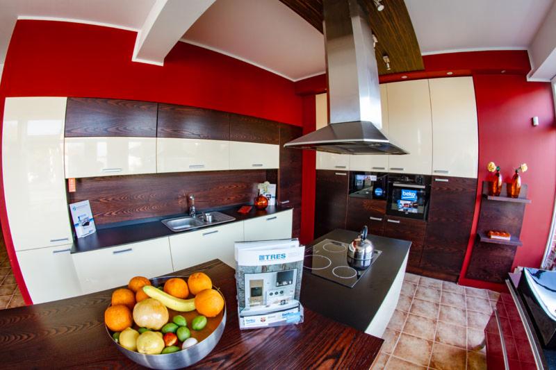 Nowoczesna kuchnia z ciemnobrązowymi i kremowymi szafkami, czerwonymi ścianami i wyspą kuchenną z czarnym blatem, na której stoi czajnik. Na pierwszym planie misa z owocami.