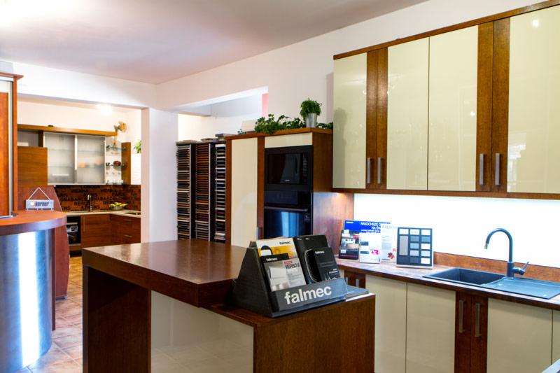 Ekspozycja mebli kuchennych w salonie: kremowe szafki z drewnianymi akcentami, wyspa z ciemnego drewna, próbki materiałów i katalogi Falmec.