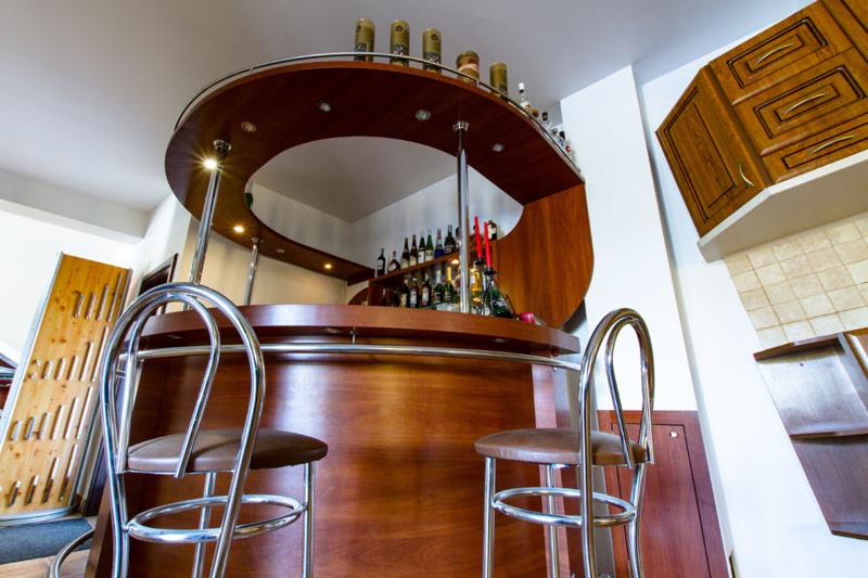 Drewniany bar z chromowanymi detalami i oświetleniem punktowym, widoczne dwa hokery z brązowymi siedziskami, w tle półki z butelkami i fragment zabudowy kuchennej.