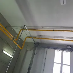 Żółte rury instalacyjne pod sufitem w hali przemysłowej, widoczne mocowania i elementy konstrukcyjne, w tle brama przemysłowa i paleta.