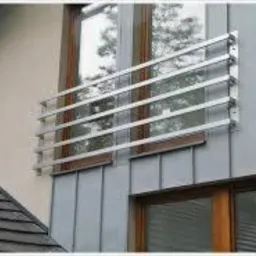 Zewnętrzny grzejnik dekoracyjny, zamontowany na elewacji budynku z drewnianą stolarką okienną i metalowym sidingiem. Grzejnik wykonany z poziomych, metalowych rurek.