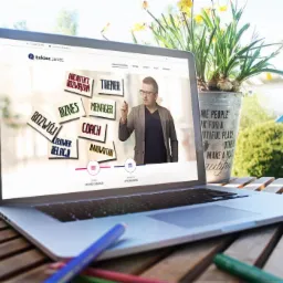 Laptop wyświetlający stronę internetową Łukasza Janiec z hasłami: Architekt Rozwiązań, Trener, Biznes, Menager, Coach, Innowator, Rozwój, Człowiek Relacji, na drewnianym stole, z doniczką...