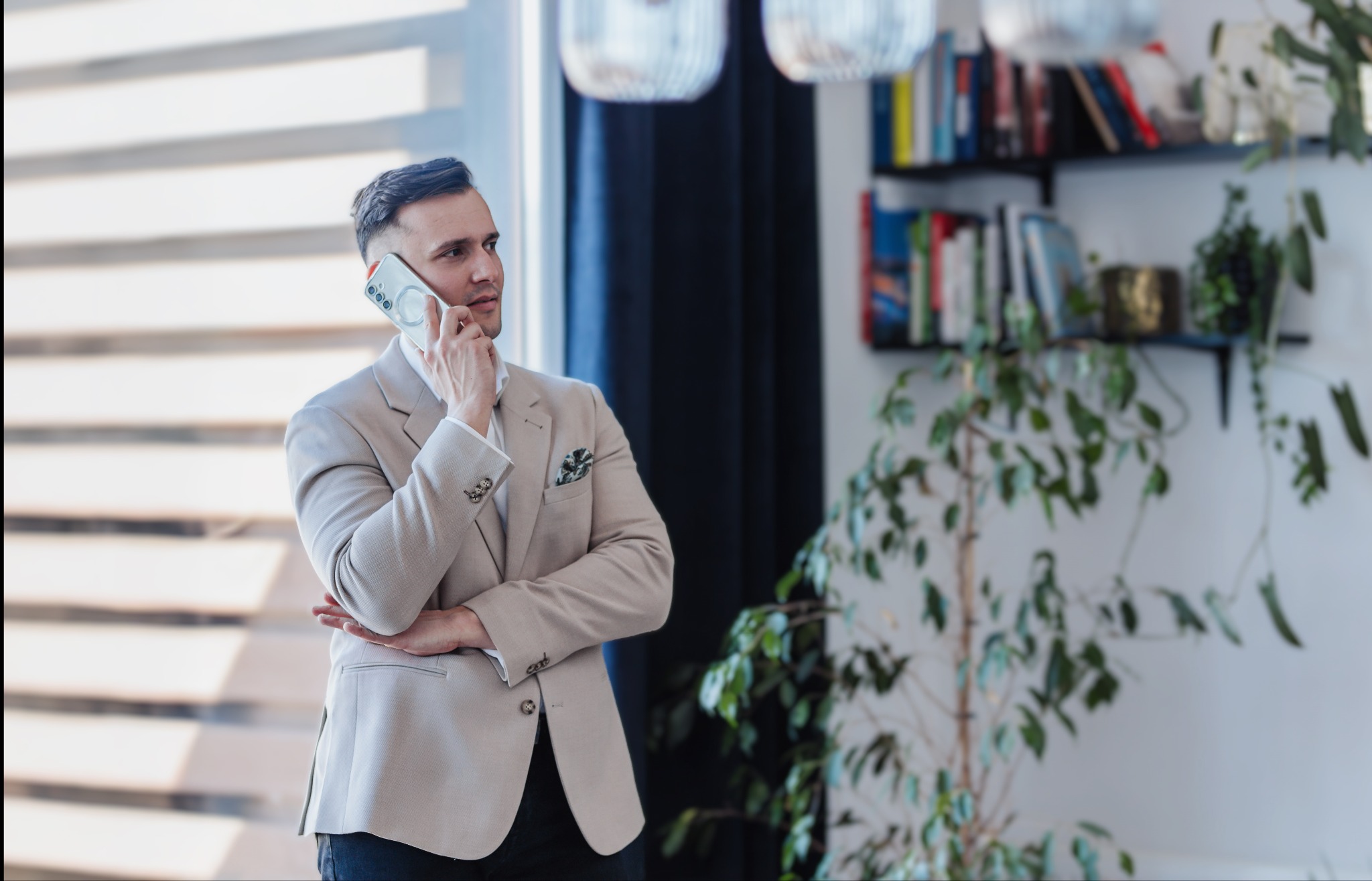 Mężczyzna w beżowej marynarce rozmawia przez telefon, stojąc na tle jasnego okna i półki z książkami oraz roślinami doniczkowymi.