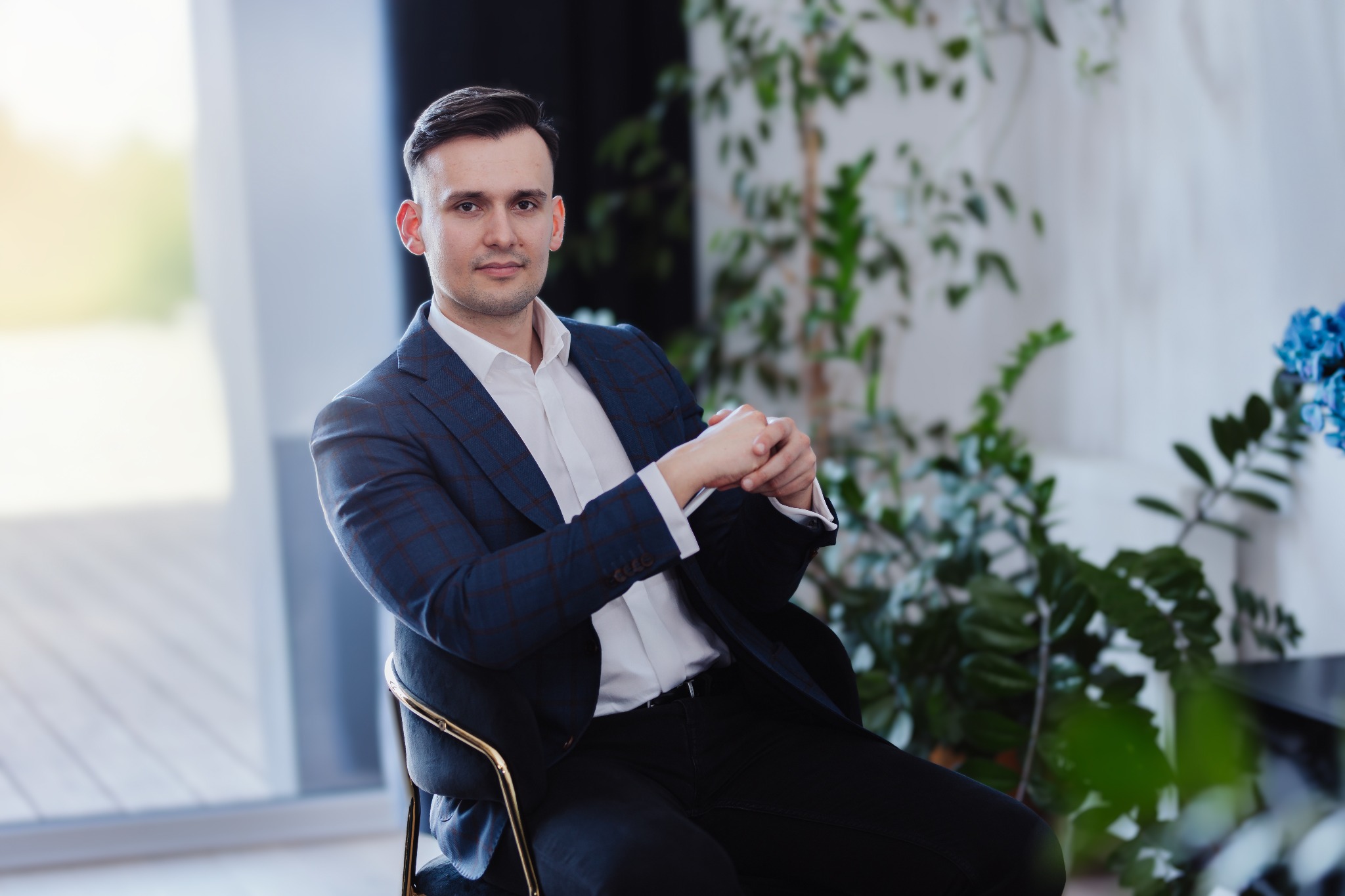 Mężczyzna w granatowym garniturze w kratę siedzi na krześle na tle roślin doniczkowych i okna.