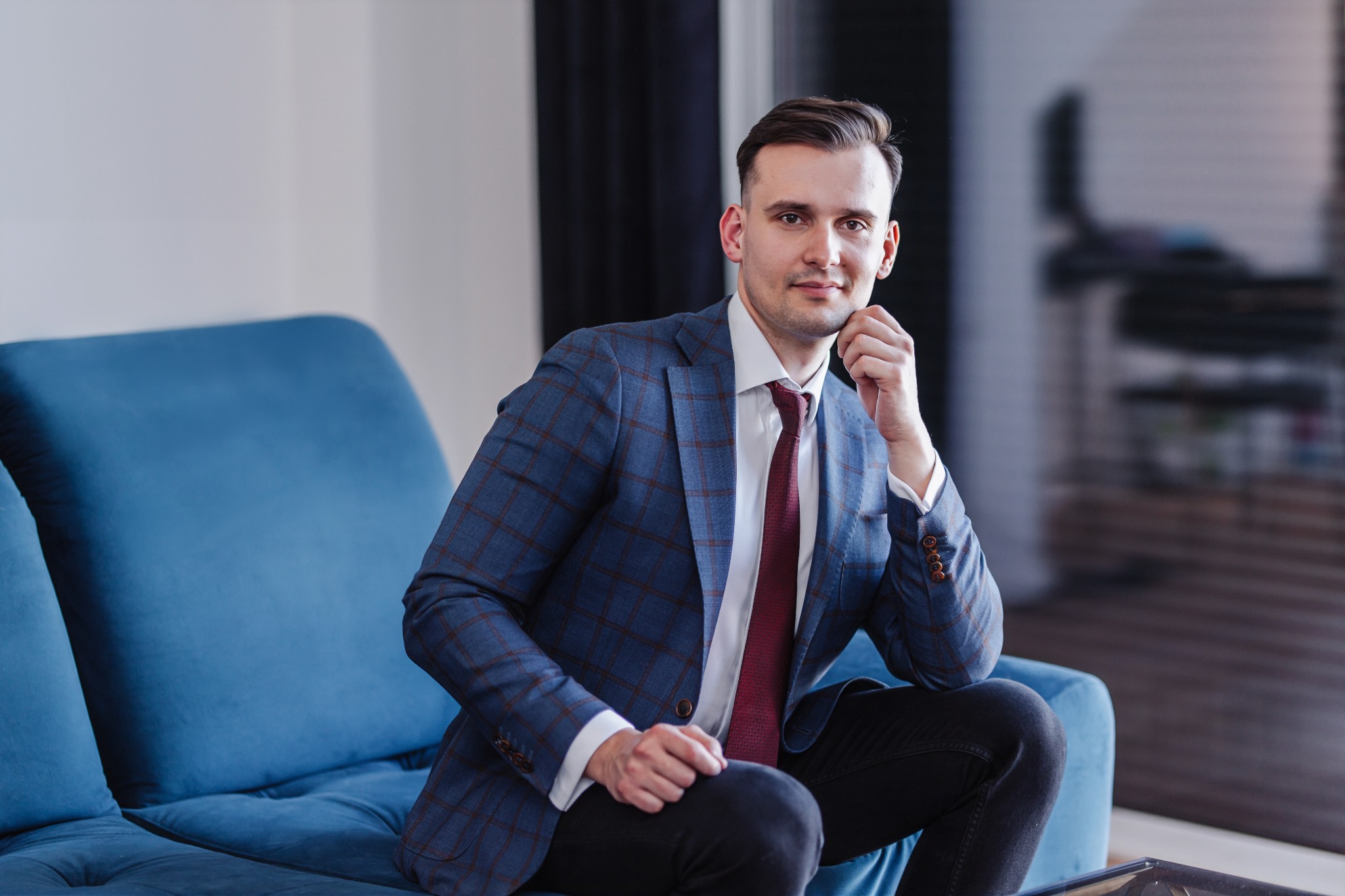 Młody mężczyzna w niebieskiej marynarce w kratę i bordowym krawacie siedzi na niebieskiej sofie z ręką podpartą pod brodą, spoglądając w obiektyw.