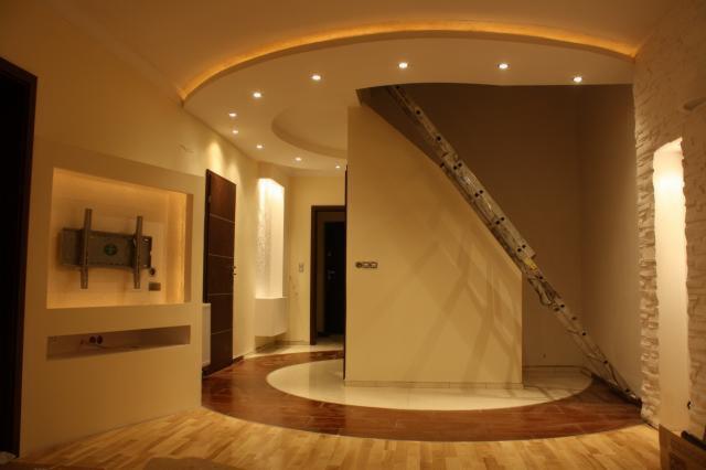 Wykończone wnętrze z podświetlanym sufitem, drewnianą podłogą z okrągłym wzorem i zamontowanym uchwytem na telewizor, widoczna drabina oparta o ścianę.