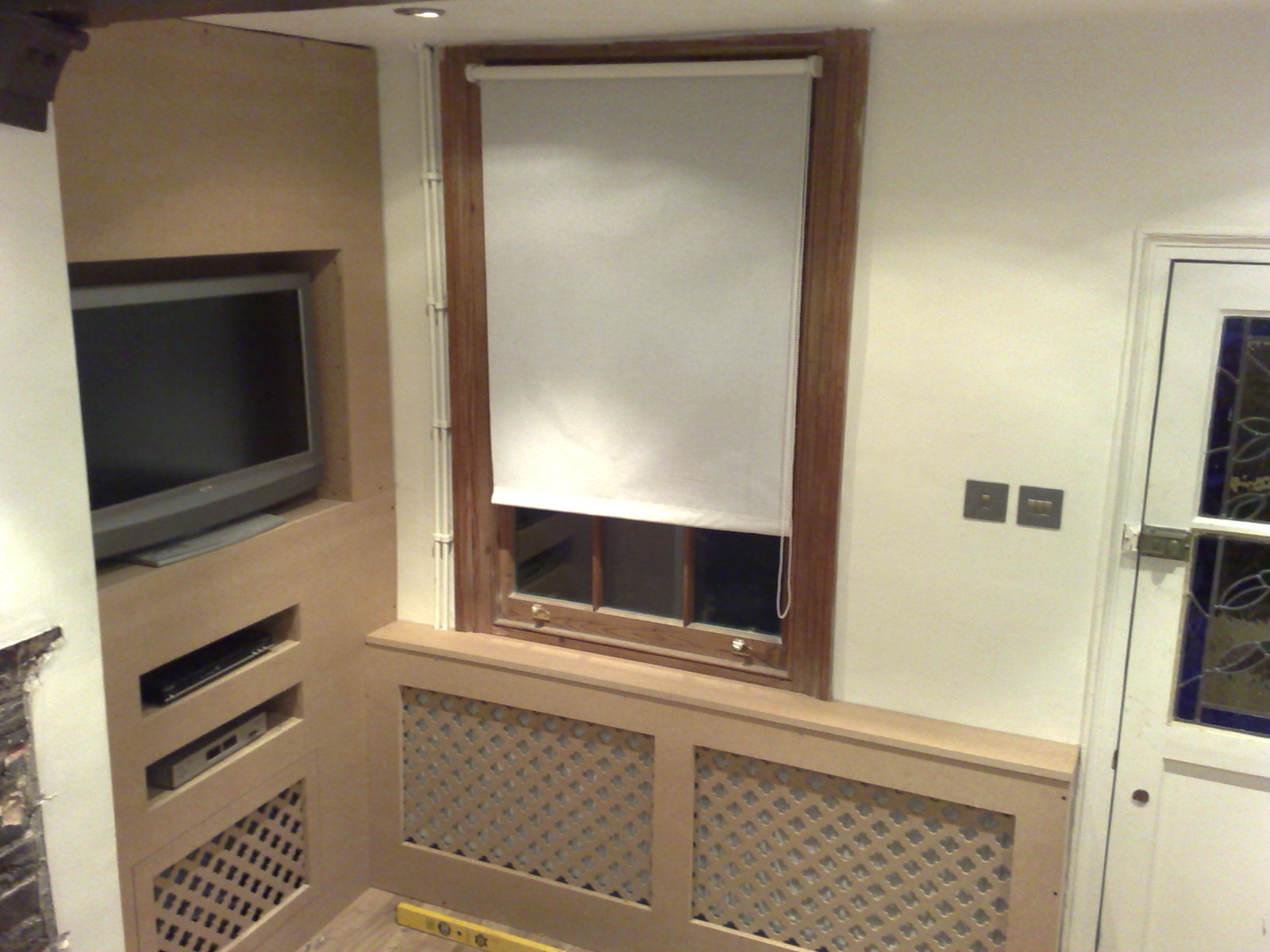 Wnęka na telewizor i sprzęt RTV w zabudowie z płyty MDF, kratka maskująca grzejnik, okno z roletą zwijaną, fragment drzwi z witrażem w trakcie prac wykończeniowych.