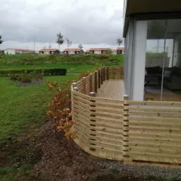 Nowoczesny taras drewniany z jasnego drewna z niską, poziomą balustradą, przylegający do budynku z dużymi oknami, widok na trawnik i zabudowę w tle, pochmurne niebo.