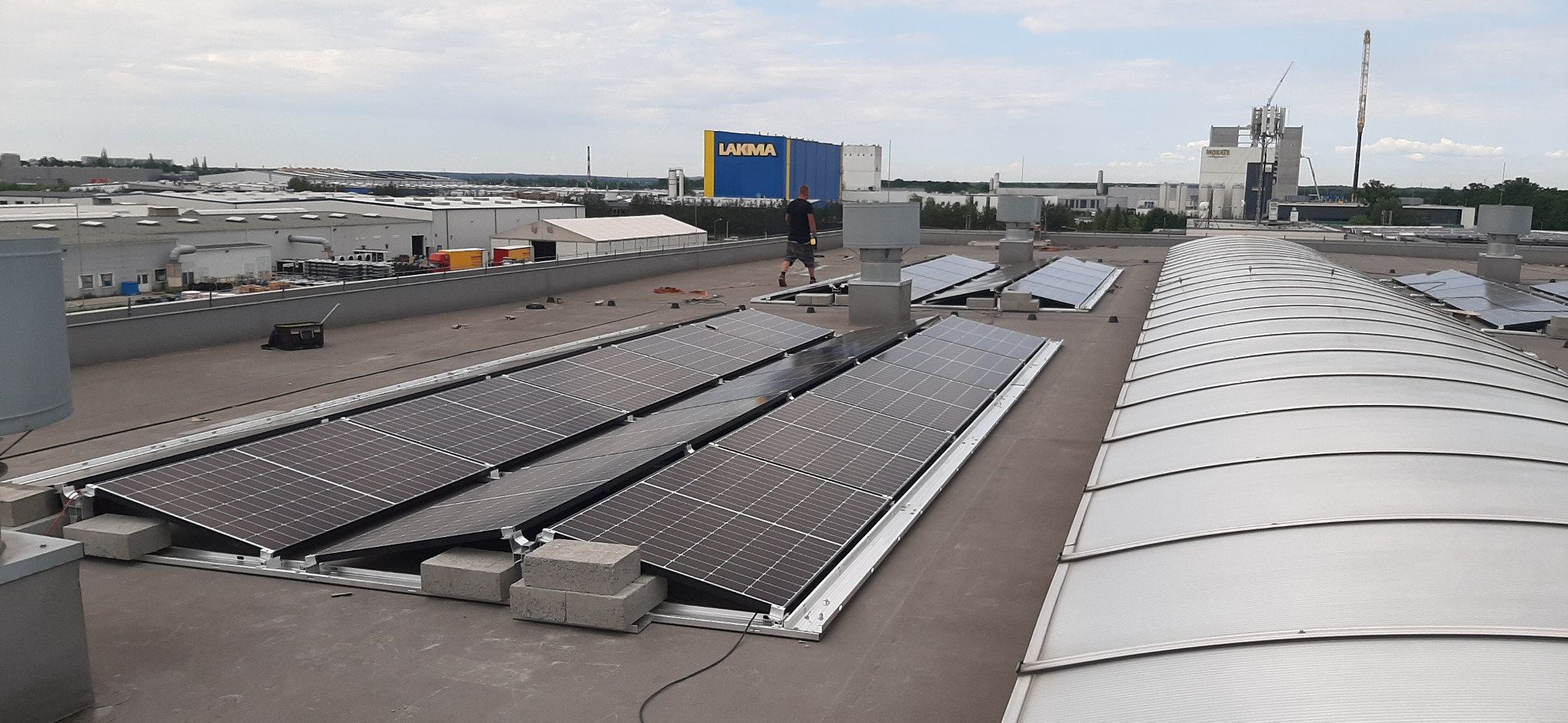 Instalacja paneli fotowoltaicznych na dachu przemysłowym z widocznym pracownikiem w tle i budynkiem Lakma.