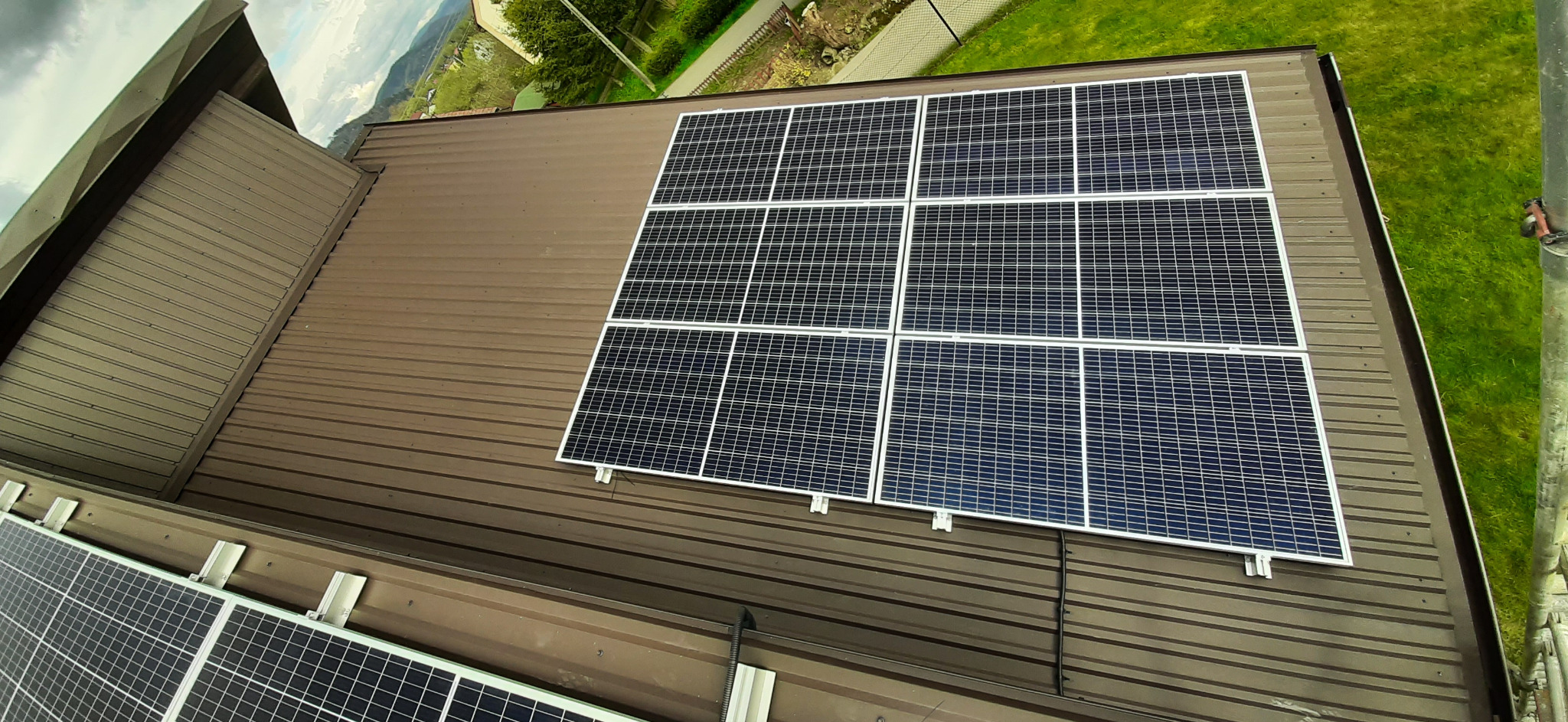 Panele słoneczne zamontowane na brązowym, metalowym dachu domu z widokiem na zielony trawnik i drzewa w tle.