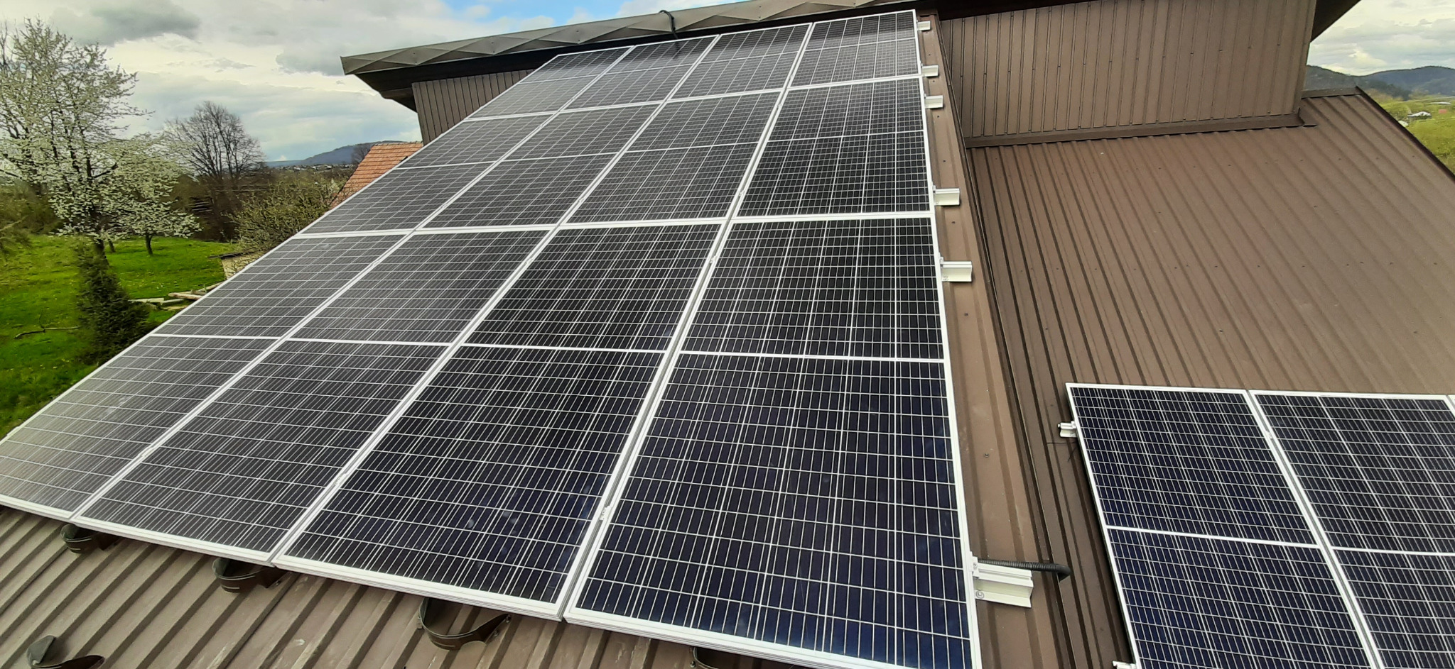 Panele fotowoltaiczne zamontowane na brązowym, metalowym dachu domu jednorodzinnego, widok na zieloną okolicę w tle.