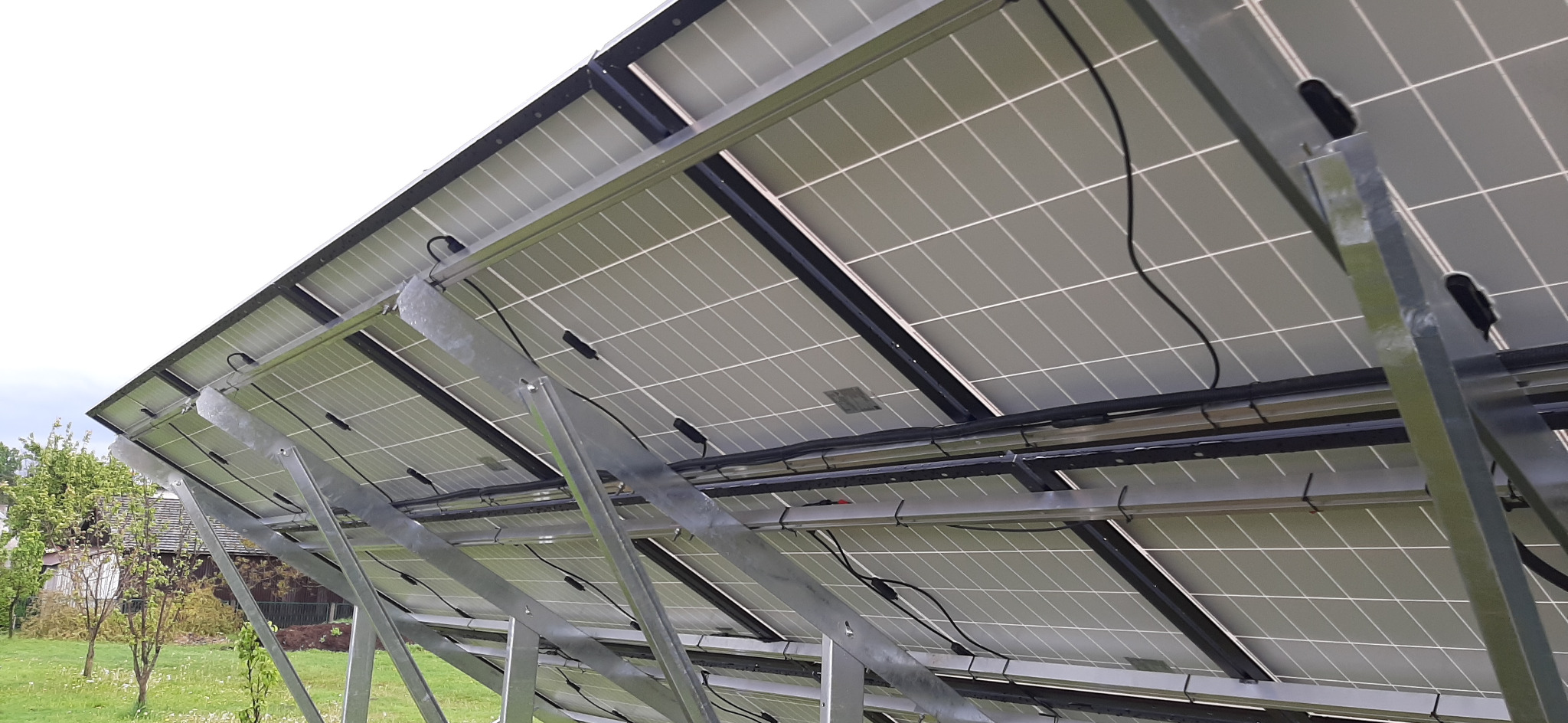 Widok od spodu na panele słoneczne zamontowane na metalowej konstrukcji, z widocznymi kablami i elementami mocującymi, na tle zielonego trawnika i domu.