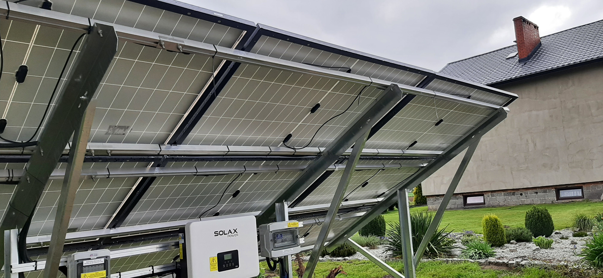 Widok od spodu paneli fotowoltaicznych zamontowanych na metalowej konstrukcji, z widocznym inwerterem Solax Power i skrzynką elektryczną, na tle budynku mieszkalnego z ceglanym kominem.