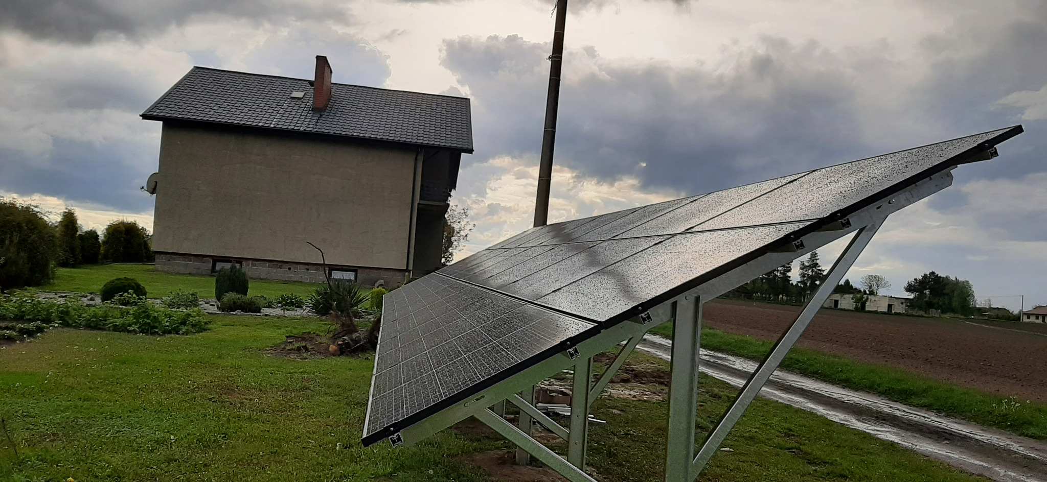 Panel słoneczny na metalowej konstrukcji, widok na dom z ceglanym kominem i pochmurne niebo