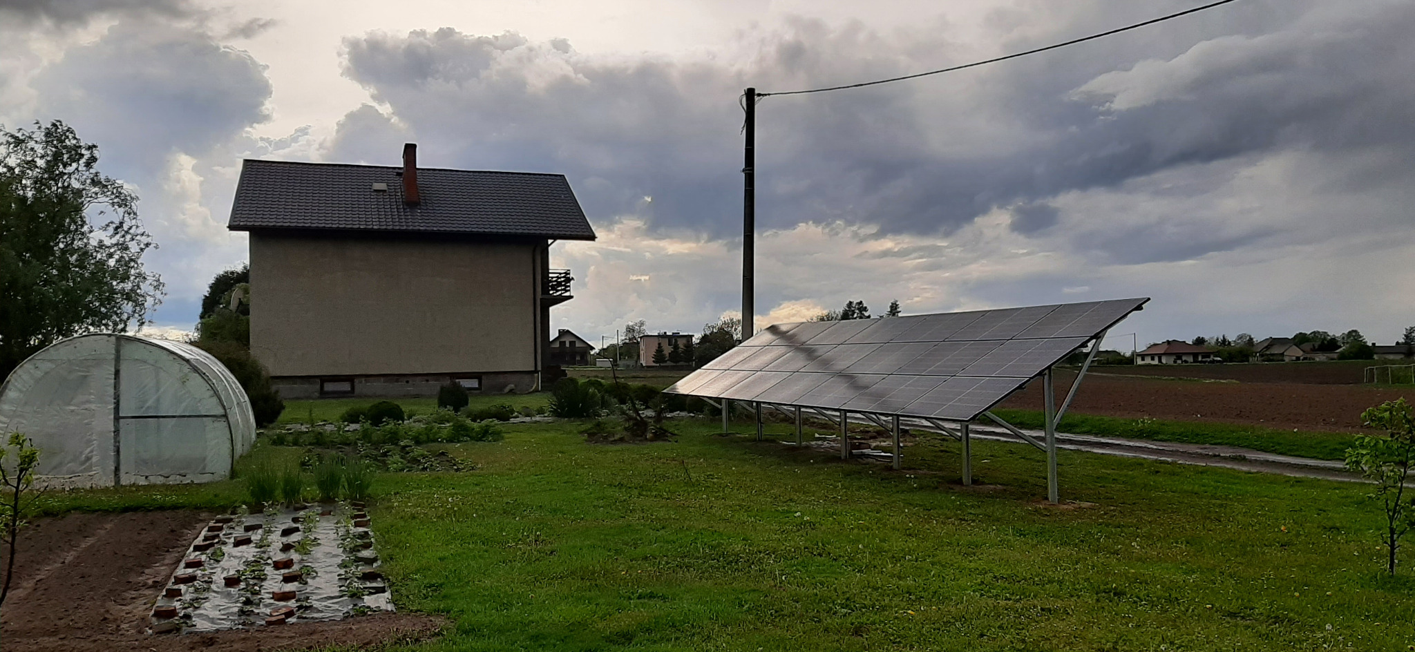 Panele słoneczne na metalowej konstrukcji, widok na dom z szarą dachówką i chmurne niebo w tle.