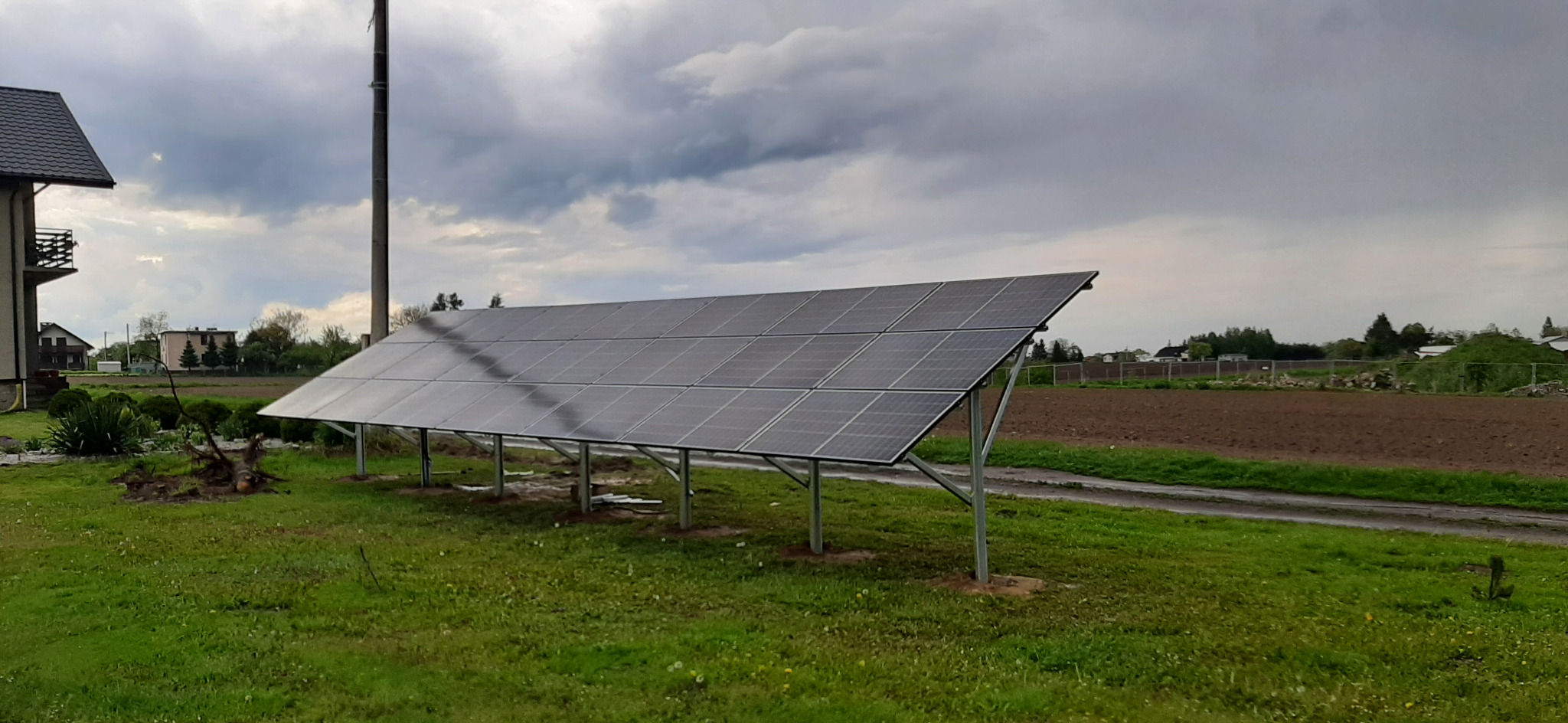 Panel fotowoltaiczny na metalowej konstrukcji, zainstalowany na trawniku przed domem, z polem w tle i pochmurnym niebem.