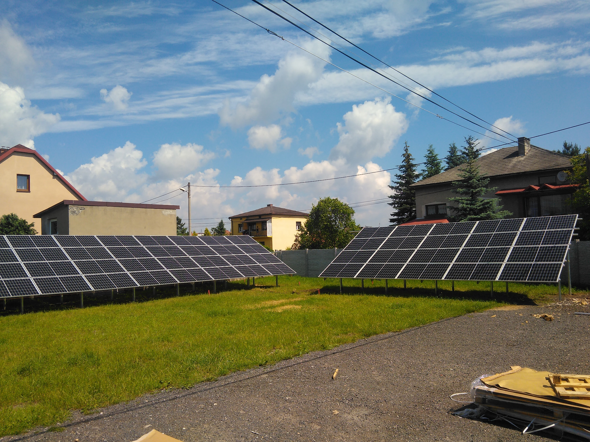 Dwa rzędy paneli fotowoltaicznych na trawie w Chorzowie, z widocznymi budynkami w tle i błękitnym niebem z chmurami.