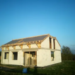 Niedokończony, dwukondygnacyjny budynek z białych bloczków, z widoczną konstrukcją dachu i otworami okiennymi, na tle zielonej trawy i błękitnego nieba.