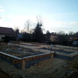 Wykopane fundamenty pod budowę domu, z szalunkiem z desek i wystającymi prętami zbrojeniowymi, widok na okoliczne budynki mieszkalne w tle.