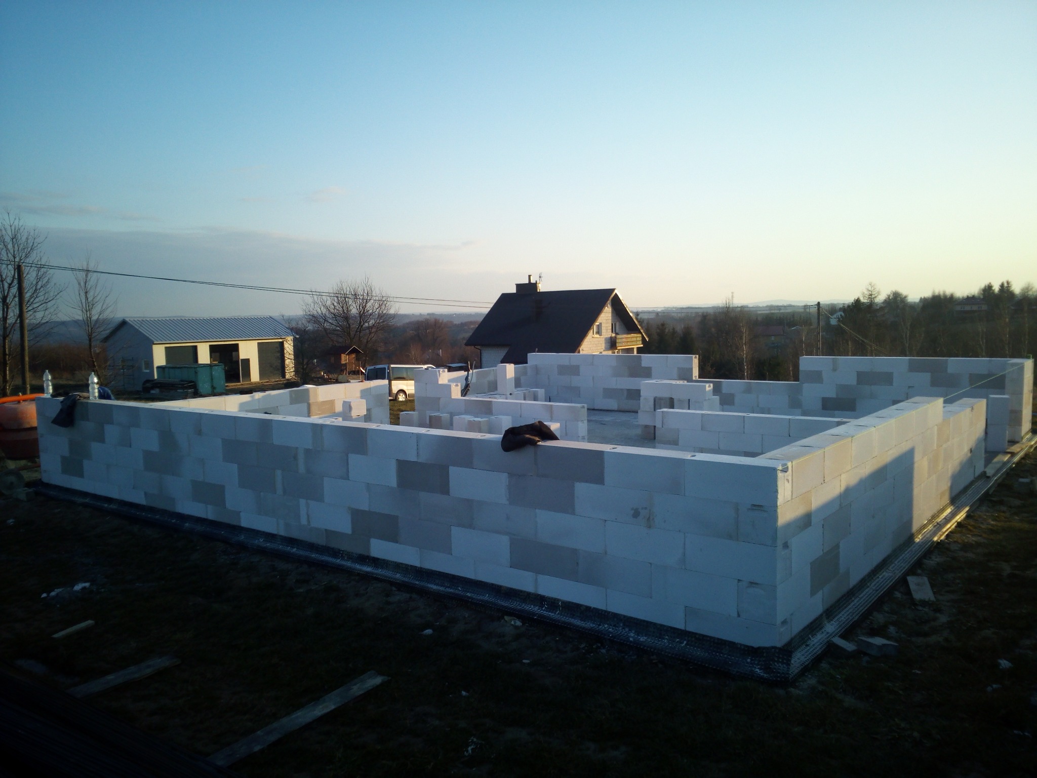 Wznoszenie ścian parteru domu z betonu komórkowego Ytong w trakcie budowy, widoczne zbrojenie fundamentów, w tle zabudowa jednorodzinna i drzewa oświetlone zachodzącym słońcem.