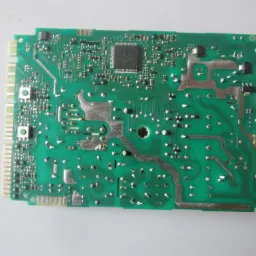 Zielona płytka drukowana programatora pralki z widocznymi elementami elektronicznymi i ścieżkami, leżąca na jasnym tle.