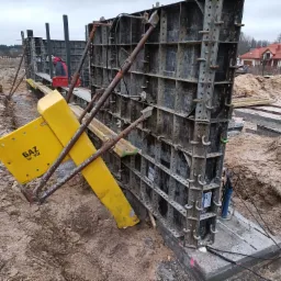 Szare, modułowe szalunki stalowe wsparte żółtymi stemplami stabilizującymi wykop, z napisem 'GAZ tel. 992', widoczne elementy zbrojenia i instalacji, w tle domy jednorodzinne.