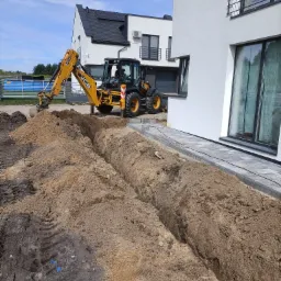 Żółta koparko-ładowarka JCB wykopuje rów w ziemi obok nowo ułożonej kostki brukowej przy nowoczesnym, białym domu z ciemną stolarką okienną i panelami słonecznymi na dachu.