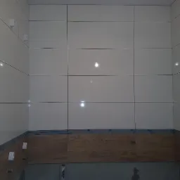 Ściana w łazience wyłożona białymi, błyszczącymi płytkami ceramicznymi w dużym formacie, z dekoracyjnym pasem płytek imitujących drewno u dołu, zabezpieczonych folią ochronną.