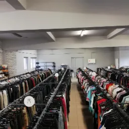 Wnętrze sklepu z odzieżą używaną; rzędy wieszaków z ubraniami, oznaczone rozmiarami, pod sufitem z widocznymi instalacjami.