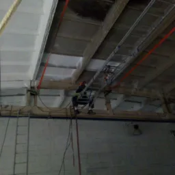 Pracownik w uprzęży, zawieszony na linach, pracuje na wysokości pod sufitem hali, używając drabiny rozstawionej na konstrukcji nośnej; widoczne elementy konstrukcji dachu i ściany.