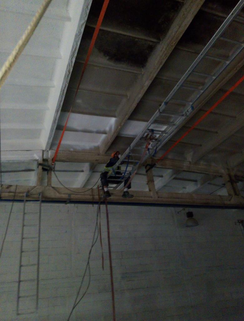 Pracownik w uprzęży, zawieszony na linach, pracuje na wysokości pod sufitem hali, używając drabiny rozstawionej na konstrukcji nośnej; widoczne elementy konstrukcji dachu i ściany.