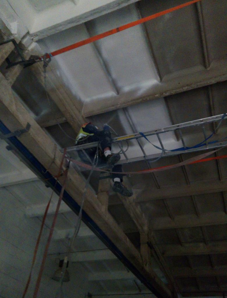 Pracownik w kasku zabezpieczony linami, balansuje na drabinie pod sufitem podczas prac na wysokościach, widoczne drewniane elementy konstrukcyjne i pomalowany sufit.