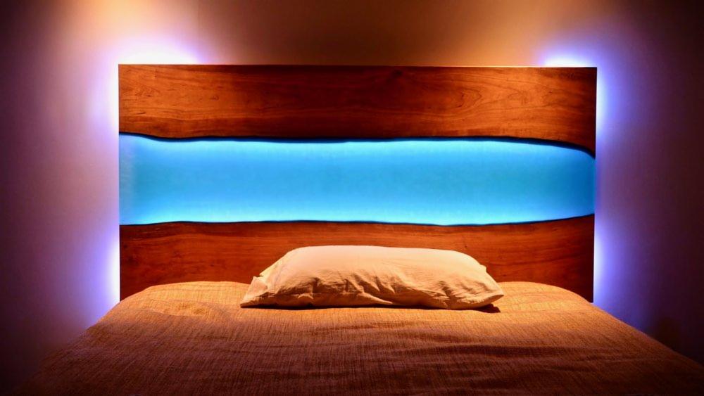 Drewniane wezgłowie łóżka z wbudowanym podświetleniem LED w kolorze turkusowym, o nieregularnym kształcie, na tle ściany z subtelnym fioletowym oświetleniem.