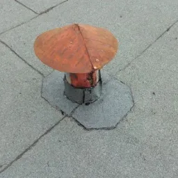 Szczegółowe ujęcie kominka wentylacyjnego z miedzianą czapką na płaskim dachu pokrytym papą, z widocznymi łączeniami papy i uszczelnieniem wokół podstawy kominka.