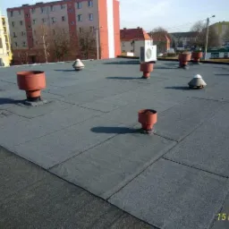 Płaski dach pokryty papą z licznymi kominkami wentylacyjnymi, niektóre w kolorze czerwonym, na tle budynków mieszkalnych w słoneczny dzień.