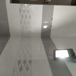 Ściana wyłożona białymi i szarymi płytkami z dekoracyjnym wzorem rombów, oświetlona lampą roboczą, widoczne otwory pod instalacje elektryczne.