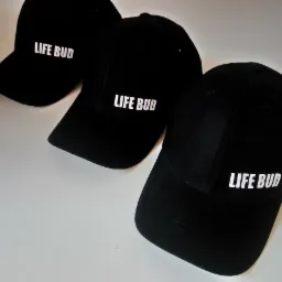 Trzy czarne czapki z daszkiem leżące na białym tle, z białym napisem 'LIFE BUD' na każdej z nich, wykonane w Warszawie.