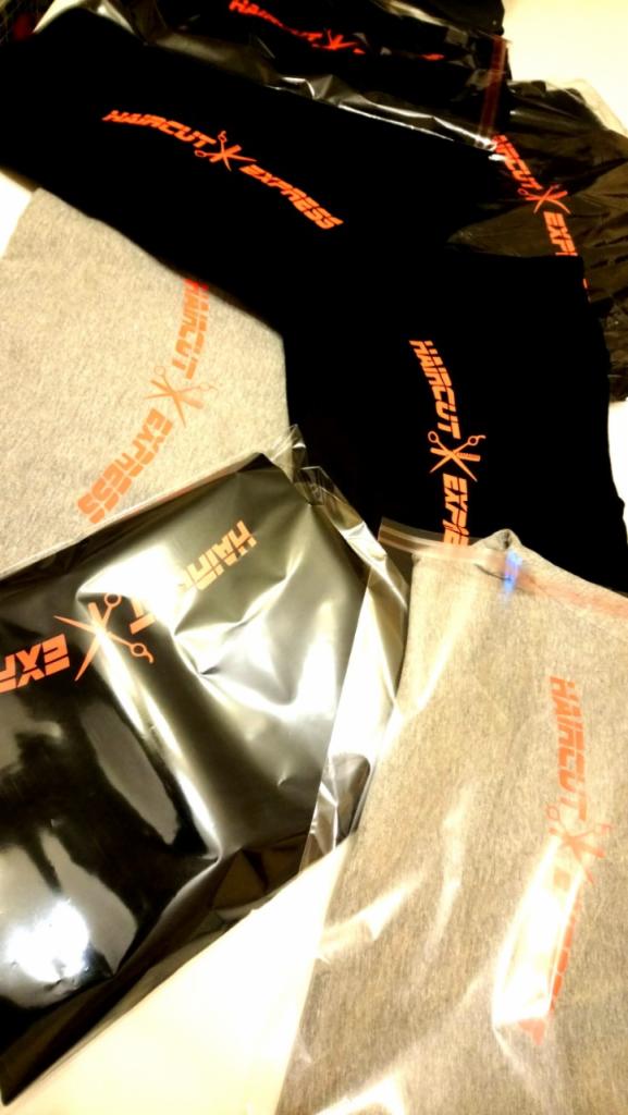 Czarne i szare koszulki z pomarańczowym napisem 'HAIRCUT EXPRESS' i symbolem nożyczek, częściowo zapakowane w przezroczystą folię.