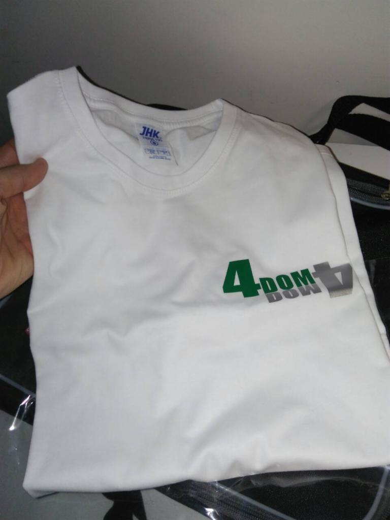Biała koszulka z krótkim rękawem, leżąca na folii, z zielonym i szarym nadrukiem '4 DOM DON' na piersi. Widoczna metka JHK przy dekolcie.